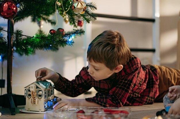 Kleine schattige blanke jongen die thuis onder de kerstboom ligt te spelen met papieren speelgoedhuis