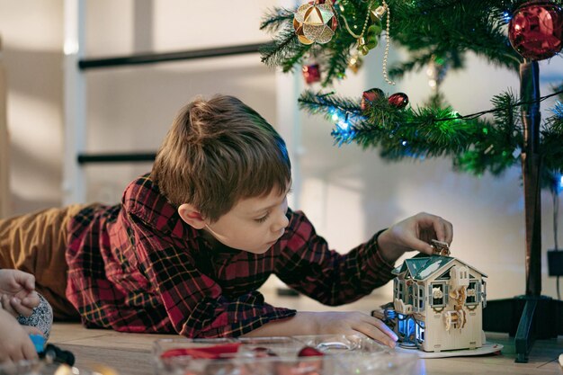 Kleine schattige blanke jongen die thuis onder de kerstboom ligt te spelen met papieren speelgoedhuis