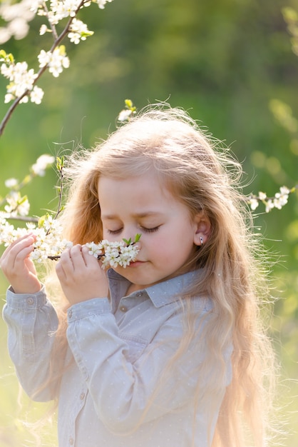 Kleine schattige blonde meid met lang haar snuift een bloeiende boomtak in het park in het voorjaar.