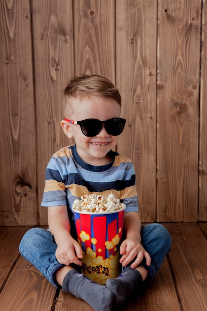 Kleine schattige jongen, 3D-bioscoopglazen met emmer voor popcorn, fastfood eten op houten