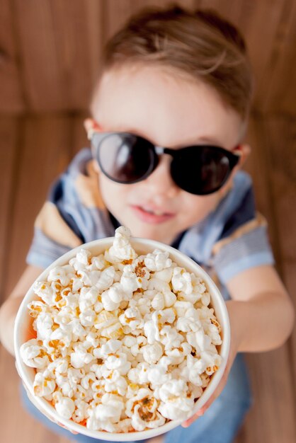 Kleine schattige jongen babyjongen 2-3 jaar oud, 3d imax bioscoop bril met emmer voor popcorn, fastfood eten op houten achtergrond.