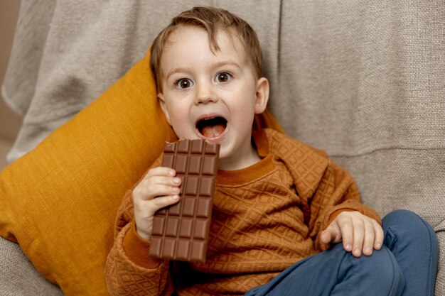 Kleine schattige jongen die thuis op de bank zit en chocoladereep eet Kind en snoep suikerwerk Kid geniet van een heerlijk dessert Kleuterkind met vrijetijdskleding Positieve emotie