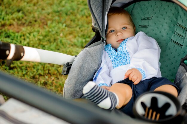 Kleine schattige jongen gekleed in het geborduurde shirt liggend in de groene kinderwagen