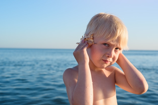 Kleine schattige jongen met zeeschelp in de hand op de achtergrond van de zee. Kind luisteren naar zee