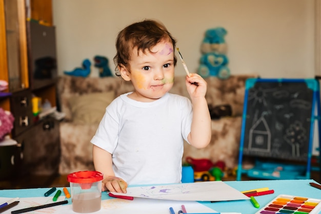 kleine schattige jongen tekent met penselen en gekleurde verf op een vel papier