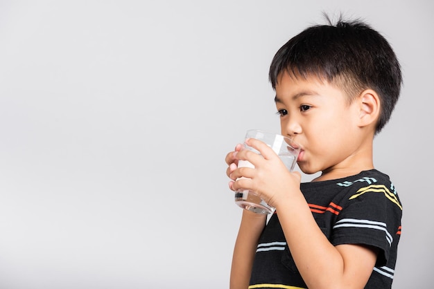 Kleine schattige jongen van 56 jaar oud glimlacht en drinkt vers water uit glas in studio-opname geïsoleerd