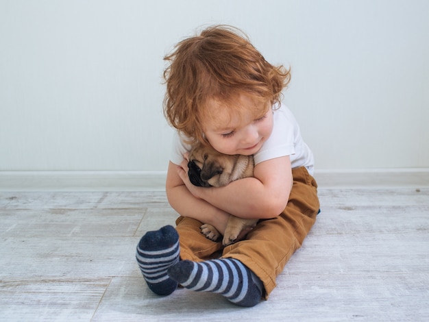 Kleine schattige roodharige krullende jongen in wit t-shirt met zijn pug puppy