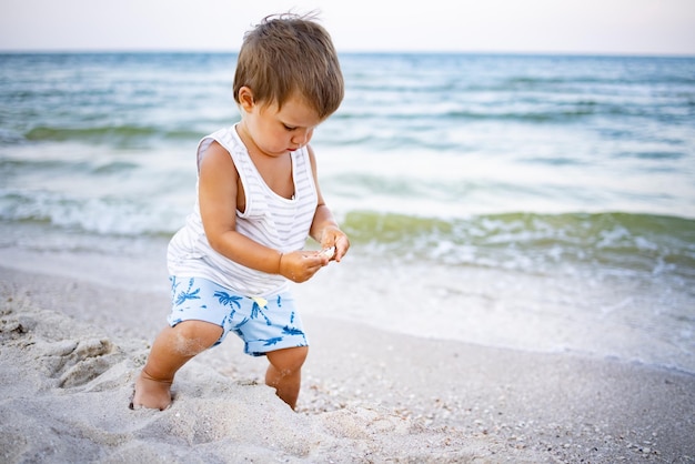 Kleine schattige vrolijke jongen in een gestreept grijswit T-shirt zoekt en verzamelt natte kiezelstenen en schelpen in het zand aan de kust van Azov op een warme zonnige zomeravond