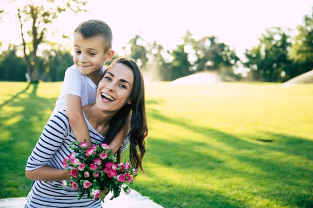 Kleine schattige zoon maakt verrassing met bloemen in handen voor zijn gelukkige en mooie jonge moeder buiten in het park