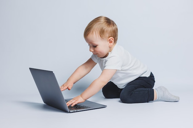 Kleine slimme jongen van 34 jaar oud zit met een laptop op een witte achtergrond een kind in een witte t-shirt en zwarte broek zit op een laptop en kijkt naar het scherm moderne progressieve kinderen