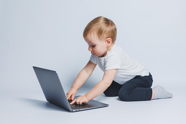 Kleine slimme jongen van 34 jaar oud zit met een laptop op een witte achtergrond Een kind in een witte T-shirt en zwarte broek zit op een laptop en kijkt naar het scherm Moderne progressieve kinderen