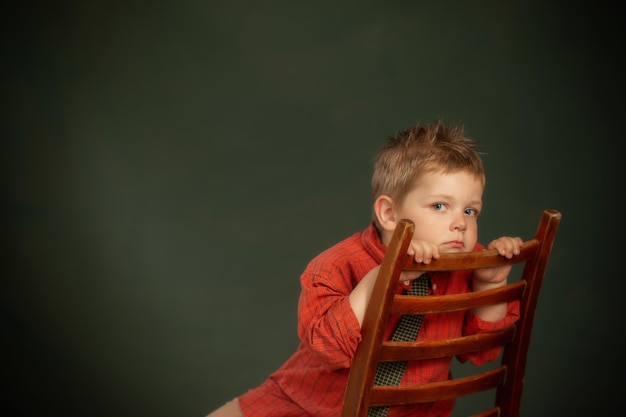 Kleine trieste jongen in rood shirt op oude stoel op donkergroene achtergrond