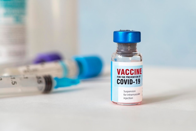 Kleine vaccinflesflesje met een etiket met de tekst Covid - 19 Corona-virusvaccin, en een medische injectiespuit met een vaccindruppel op de naald, op blauwe achtergrond