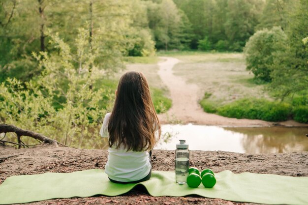 Foto kleine vrouwelijke sporter zit op een mat op de heuvel voor het meer en geniet van de natuur dumbbells en fles liggen bij het kind