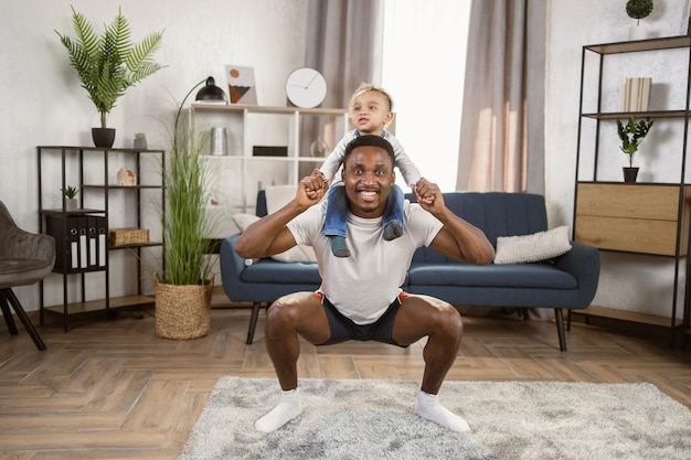 Kleine zoon zit op sterke vaderschouders en doet squat-oefeningen