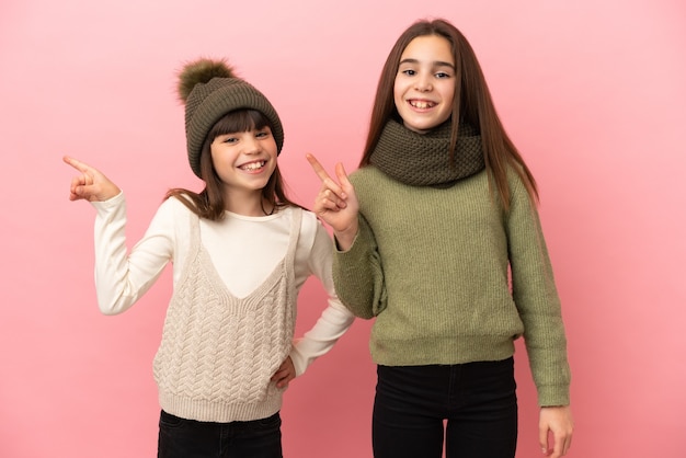 Kleine zusjes die een winterkleren dragen geïsoleerd op een roze achtergrond die met de vinger naar de zijkant wijzen en een product presenteren