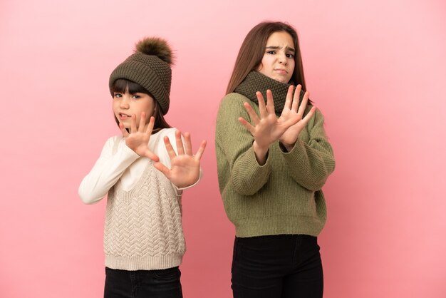 Kleine zusjes die winterkleren dragen die op een roze achtergrond zijn geïsoleerd, zijn een beetje nerveus en bang om hun handen naar voren te strekken