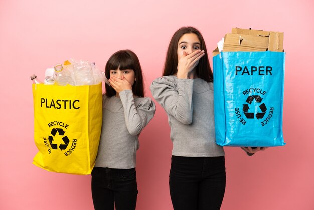 Kleine zusjes recyclen papier en plastic geïsoleerd op een roze achtergrond die de mond bedekken met handen om iets ongepasts te zeggen