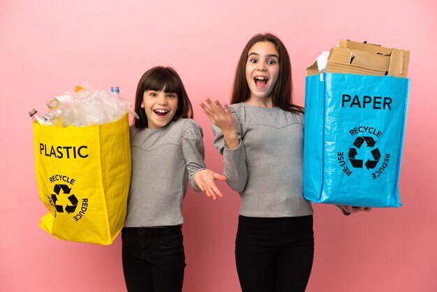 Kleine zusjes recyclen papier en plastic geïsoleerd op roze achtergrond met verrassing en geschokte gezichtsuitdrukking