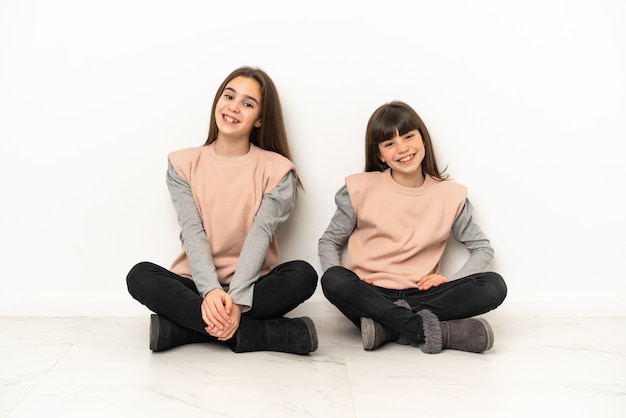Kleine zusjes zittend op de vloer geïsoleerd op een witte achtergrond poseren met armen op heup en glimlachen