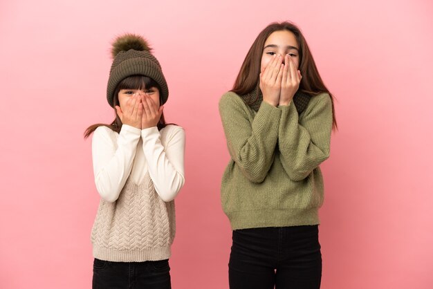 Kleine zusters die de winterkleren dragen die op roze achtergrond worden geïsoleerd die veel glimlachen terwijl zij mond bedekken