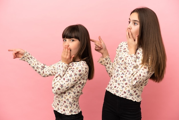 Kleine zusters meisjes geïsoleerd op roze achtergrond wijzende vinger naar de zijkant met een verbaasd gezicht