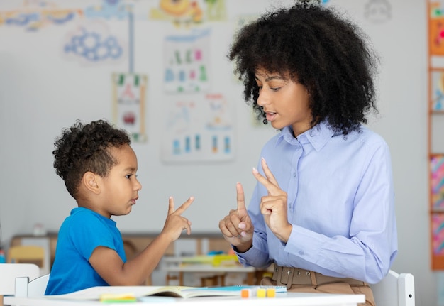 Kleine zwarte jongen leert tellen op vingers met vrouwelijke leraar hulp zitten aan het bureau in de klas