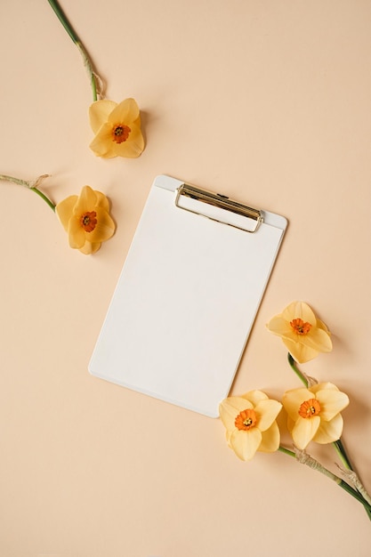 Klembord tablet pad met blanco vel papier op neutrale beige tafel Esthetische kunstenaar thuiskantoor bureau werkruimte met narcissen bloemen Plat lag bovenaanzicht mockup met lege kopie ruimte