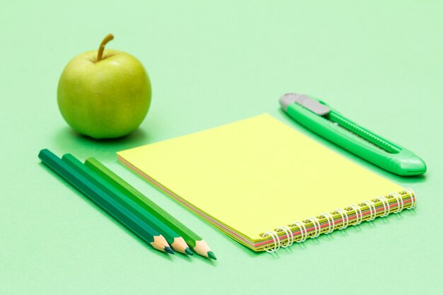 Kleurpotloden, notitieboekje, papiermes en appel op groene achtergrond. terug naar schoolconcept. schoolspullen. ondiepe scherptediepte.