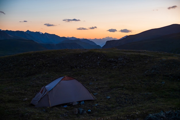 Kleurrijk dageraadlandschap met tent op pas tegen lila bergen onder oranje dageraadhemel met violette wolken. Tent op heuvel met uitzicht op grote bergen silhouetten in zonsondergang. Mooie alpiene zonsopgang.