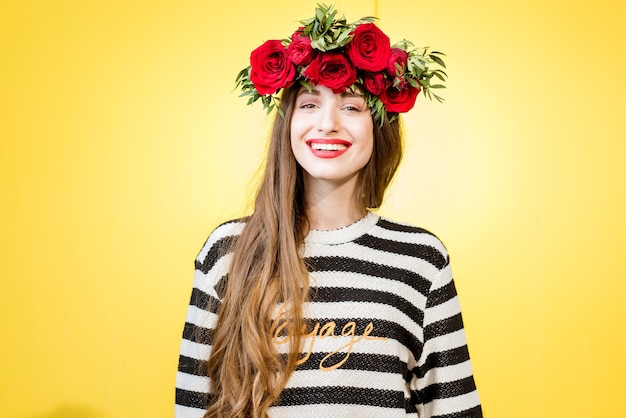 Kleurrijk portret van een mooie vrouw in trui met krans gemaakt van rode rozen op de gele achtergrond