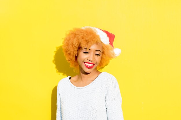 Kleurrijk portret van een schattige Afrikaanse vrouw in kerstmuts op de gele achtergrond. Winter vakantie concept