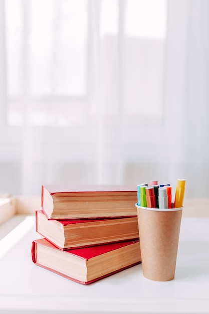 Kleurrijke markeerstiften in de ambachtelijke papieren pot op witte tafel. schoolspullen. grote rode boeken op tafel.