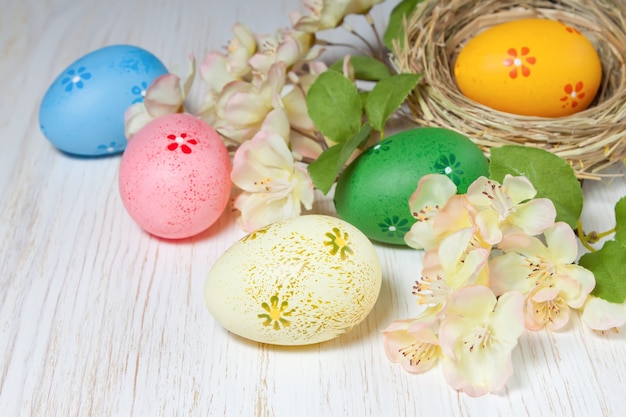 Kleurrijke paaseieren in stro nest en tak met bloemen op witte houten achtergrond