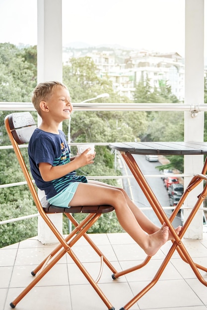 Kleuterjongen zit melk te drinken en geniet van het leven lachend zittend op een stoel op het balkon van het hotel