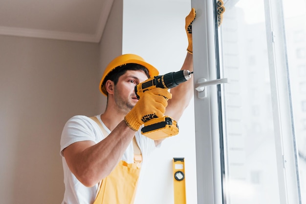 Klusjesman in geel uniform installeert nieuw raam met behulp van automatische schroevendraaier Huisrenovatieconcept