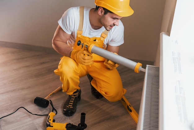 Klusjesman in geel uniform werkt binnenshuis met warmtebatterij met behulp van speciaal gereedschap Huisrenovatieconcept