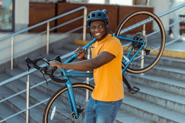 knap glimlachende Afro-Amerikaanse man met een helm die zijn fiets vasthoudt die op een stedelijke straat loopt