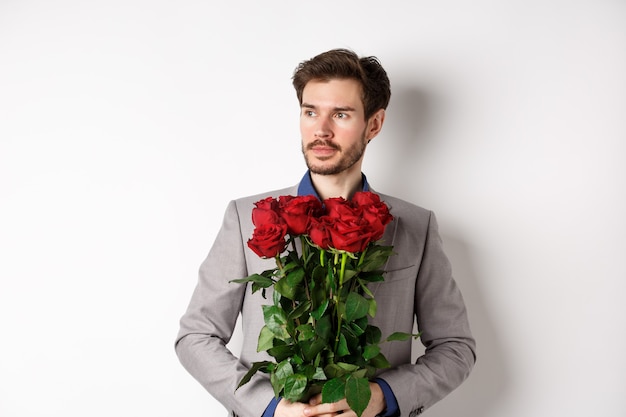 Knap vriendje in pak gaande op een romantische date, boeket van rode rozen te houden en op zoek naar links attent, staande op een witte achtergrond.