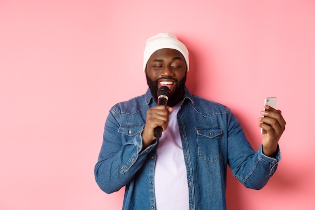 Knappe Afro-Amerikaanse man die karaoke zingt, songteksten leest op smartphone-app en microfoon vasthoudt, staande over roze achtergrond