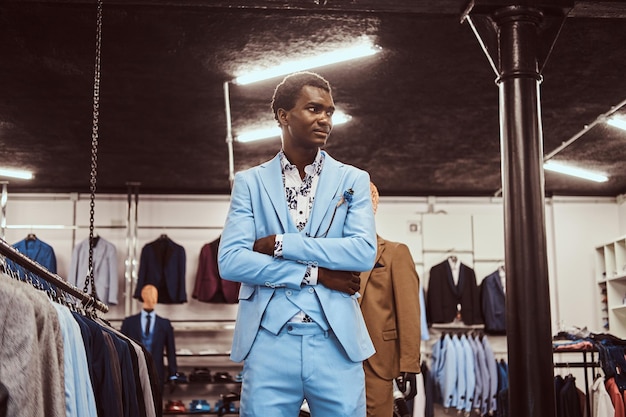 Knappe Afro-Amerikaanse man gekleed in blauw elegant pak poseren met gekruiste armen in een klassieke herenkledingwinkel.