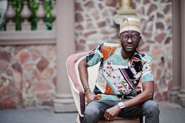 Knappe Afro-Amerikaanse man met traditionele kledingpet en bril in moderne stad zittend op een stoel