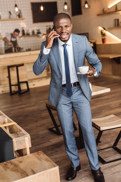 Knappe Afro-Amerikaanse zakenman kopje koffie houden en praten op smartphone in café