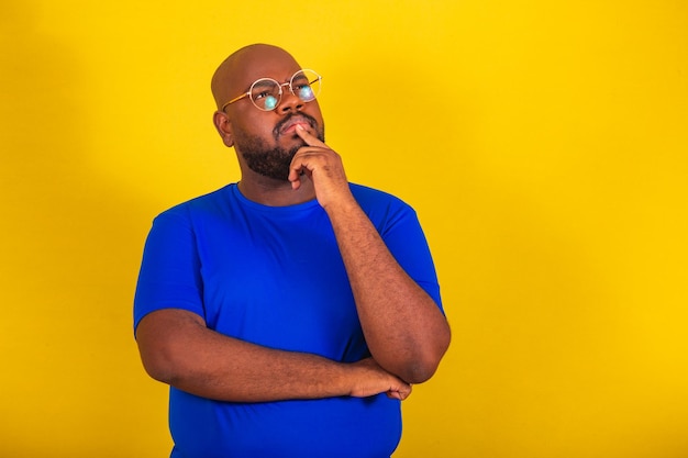 Knappe Afro-Braziliaanse man met een bril blauw shirt over gele achtergrond met handen op de kin vragende reflectie denken denken kiezen redenering