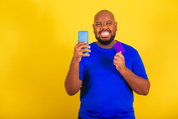 Knappe Afro-Braziliaanse man met een bril blauw shirt over gele achtergrond met mobiele telefoon en creditcard online winkelconcept inapp-aankopen die het scherm tonen