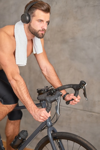 Knappe atletische man met handdoek om zijn nek luisteren naar muziek en hometrainer rijden tegen marmeren muur