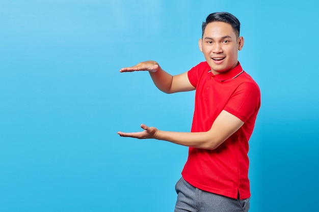 Knappe Aziatische jongeman in rood shirt glimlachend met product met palm, met kopieerruimte voor bepaalde korting geïsoleerd op blauwe achtergrond