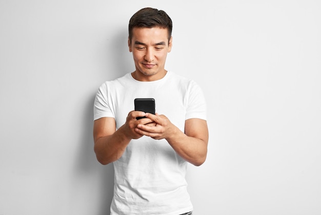 Knappe Aziatische man in witte studio glimlacht vreugdevol kijkt naar het scherm van de mobiele telefoon. Gelukkig tevreden man gekleed casual schrijft en leest sms op smartphone