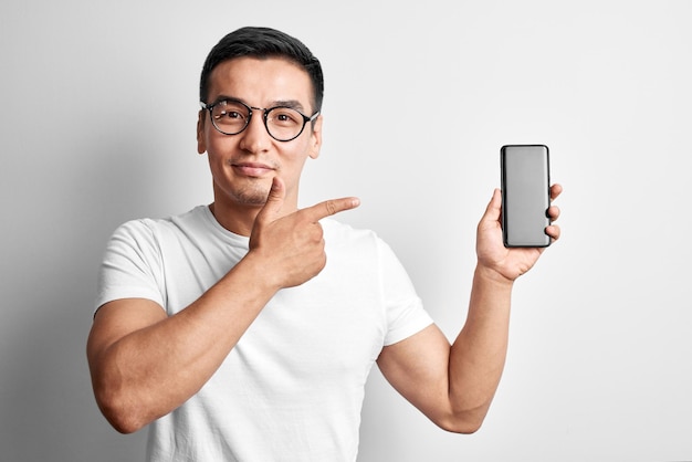 Knappe Aziatische man in witte studio glimlacht vreugdevol wijzende wijsvinger naar het scherm van de mobiele telefoon. Gelukkig man in bril gekleed casual grepen en adverteert smartphone