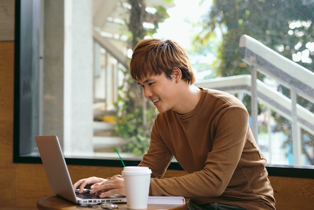 Knappe aziatische man met laptop zittend op houten tafel in coffeeshop met een kopje koffie